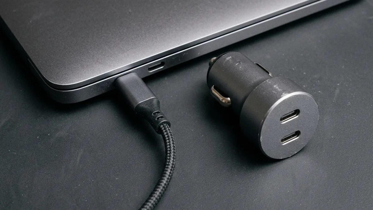 The brushed-aluminum side edge of a closed unbranded laptop showing two oval USB-C ports with one braided USB-C cable plugged in, beside an unbranded cylindrical car charger on a gray workbench mat.